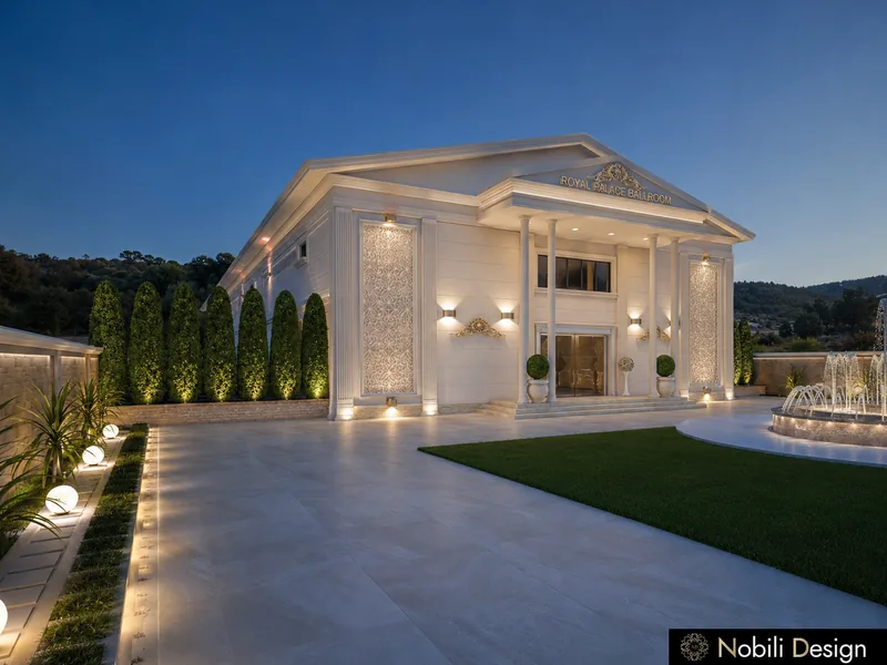 Wedding venue exterior with lit columned facade, cypress hedge, stone paving, lawn and fountain at dusk